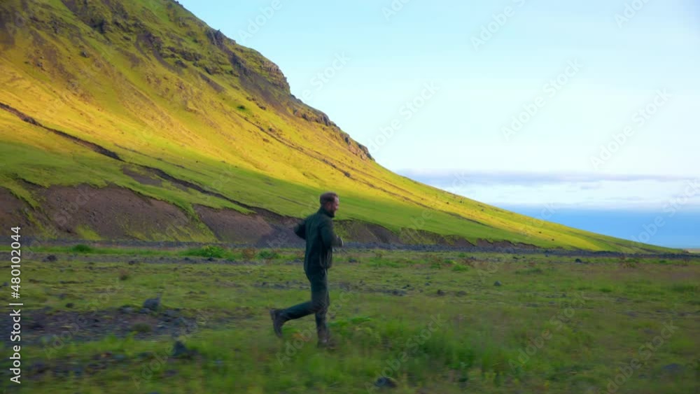 Caucasian Man Running On Grassy Terrain Near The Mountains Of Seljavallalaug In Southern Iceland. Tracking Caucasian Man Running On Grassy Terrain Near The Mountains Of Seljavallalaug In Southern Iceland. Tracking