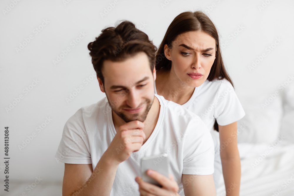 Suspicious Wife Reading Husband's Messages On Smartphone Behind Back Indoors Suspicious Wife Reading Husband's Messages On Smartphone Behind Back Indoors
