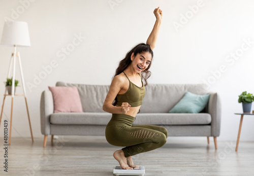 Fotografie Triumphant Indian woman in sports clothes sitting on scales, gesturing YES, sati