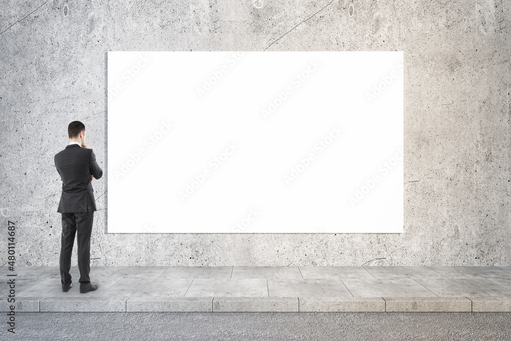 Thoughtful businessman looking at empty white poster in concrete interior. Mock up. Thoughtful businessman looking at empty white poster in concrete interior. Mock up.