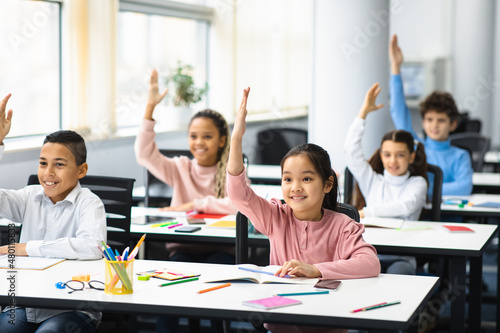 Diverse group of small schoolchildren raising hands at classroom