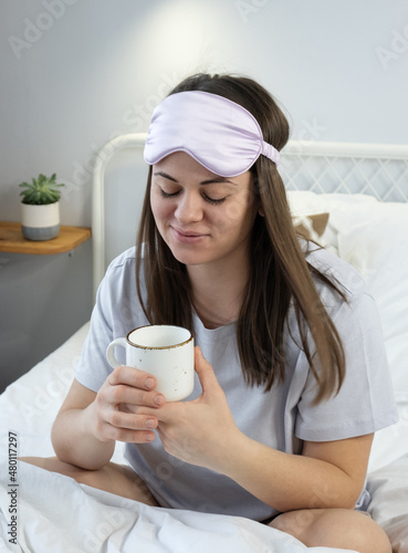 Attractive woman in silk mask having a tea on bed. Portrait of girl enjoying her drink while relaxing at home