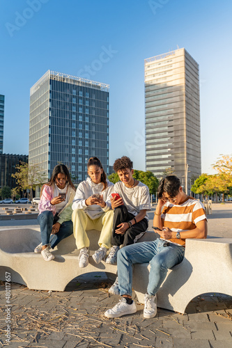 Group of happy teenage friends looking the phones in a bench in the city street.