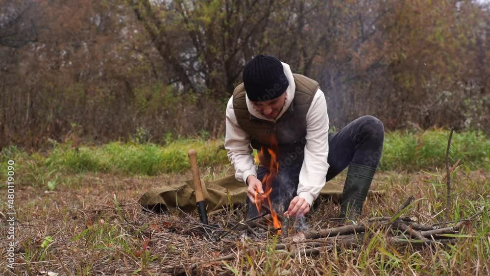 Survivalist man in warm clothes putting kindling on burning fire to keep warm and cook food at outdoors on cloudy cold day. Concept of survival, travel, hiking, camping. Shooting in slow motion. Survivalist man in warm clothes putting kindling on burning fire to keep warm and cook food at outdoors on cloudy cold day. Concept of survival, travel, hiking, camping. Shooting in slow motion.