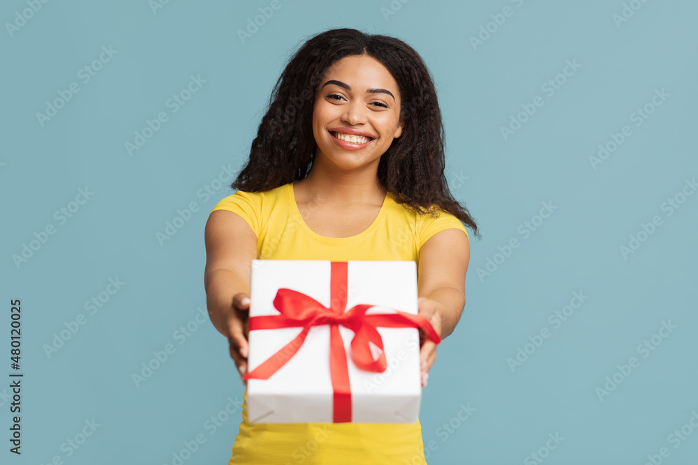 Greeting and celebration. Cheerful black lady holding wrapped gift box, stretching present to camera, blue background Greeting and celebration. Cheerful black lady holding wrapped gift box, stretching present to camera, blue background