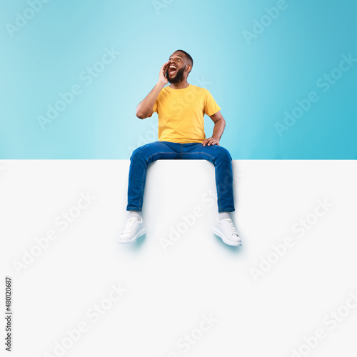 Happy black man sitting on white blank placard, shouting, announcing huge sale on blue background, mockup