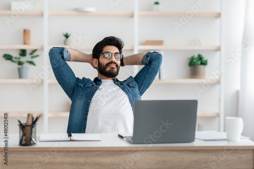 Successful arab freelancer guy resting holding hands behind head, relaxing at workplace after online work photo