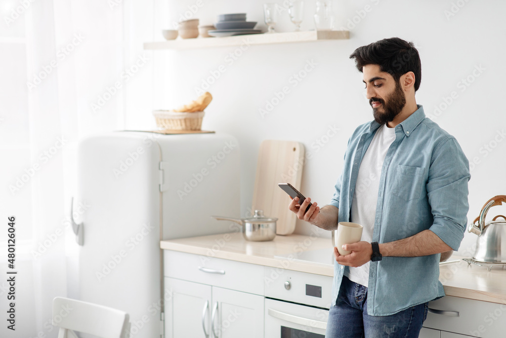 Morning routine. Young arab man using smartphone and drinking coffee in kitchen, free space