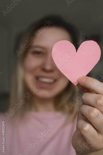 A young woman in a pink T-shirt holds a pink heart in her hand at the level of her face and smiles. A gift greeting for a holiday. Greeting card for Valentine s Day