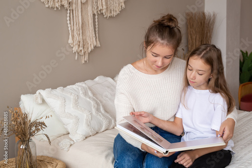 mother and daughter are sitting at home on the bed flipping through and looking at a book with photos. important moments of life in a photo album. Cute sisters read an interesting fairy tale together