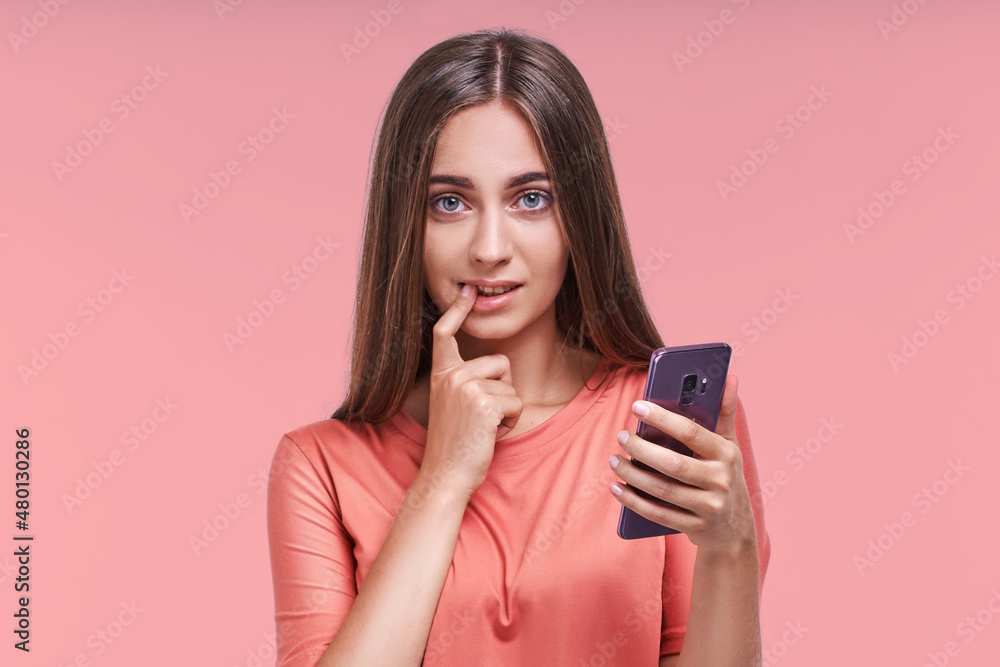 Attractive young long haired caucasian woman holds mobile phone in hand and puts finger in mouth, thinking about what to write to boyfriend flirting by SMS in messenger stands in pink studio. Attractive young long haired caucasian woman holds mobile phone in hand and puts finger in mouth, thinking about what to write to boyfriend flirting by SMS in messenger stands in pink studio.
