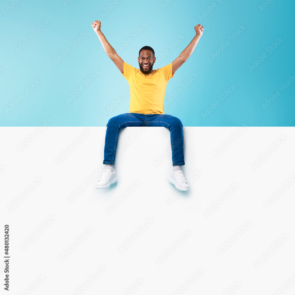 Advertising billboard template. Young black guy sitting on top of blank white poster with mockup, lifting hands up