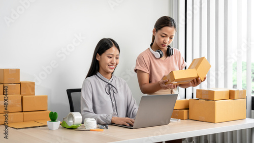 Asian woman colleague is checking customer order on laptop together and preparing packing products in parcel boxes