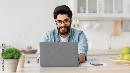 Remote business. Happy arab freelancer man working with laptop in kitchen, typing on computer, enjoying online job