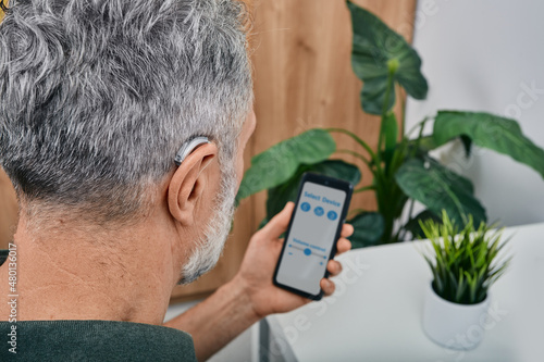 Hearing impaired mature man adjusts settings for his BTE hearing aid via smartphone. Hearing aids, deafness treatment, innovative technologies at audiology photo