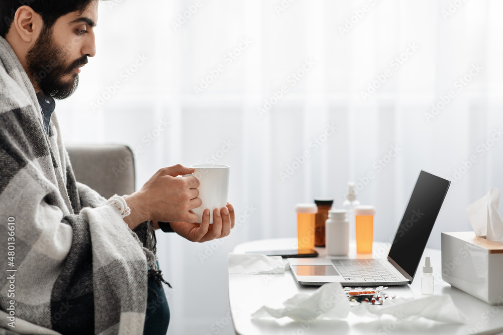 Treating flu and colds at home. Arab guy having fever wrapped in blanket and drinking hot tea, watching film on laptop Treating flu and colds at home. Arab guy having fever wrapped in blanket and drinking hot tea, watching film on laptop