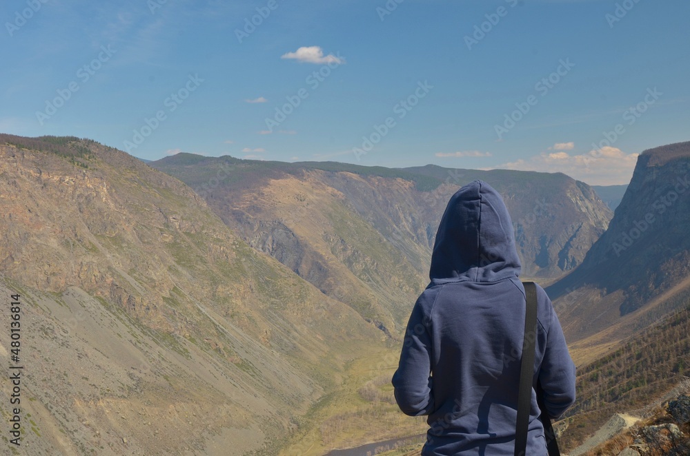 A girl in sunglasses in a blue hoodie stands against the background of a mountain valley with a blurred background in spring in sunny weather.