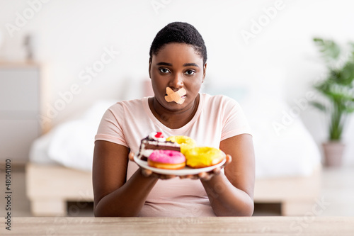 Hungry plus size black woman having weight loss diet  wearing adhesive bandages on mouth  holding plate with sweets
