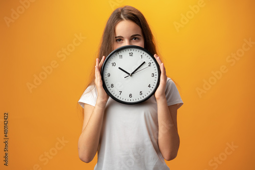 Beautiful teen girl holding wall clock over yellow background