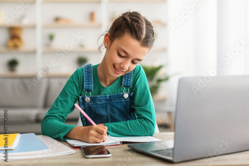Smiling pupil sitting at table, using laptop, writing in notebook