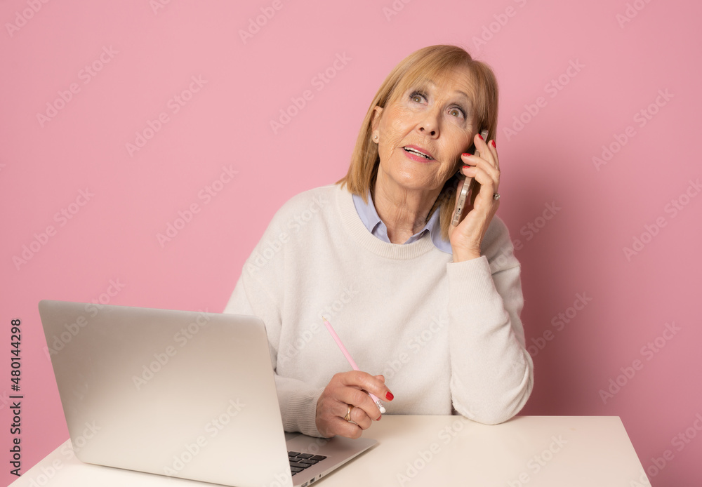 Attractive senior female sitting isolated on desk with computer talking on smartphone. Attractive senior female sitting isolated on desk with computer talking on smartphone.