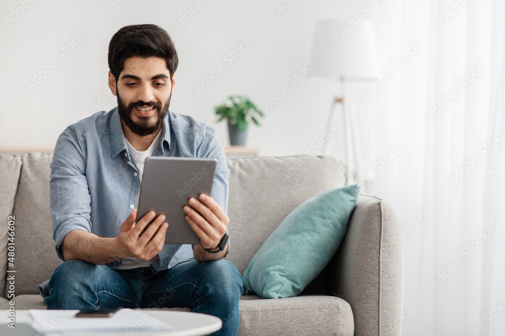 Free time concept. Portrait of happy guy using digital tablet sitting on the sofa at home, free space Free time concept. Portrait of happy guy using digital tablet sitting on the sofa at home, free space