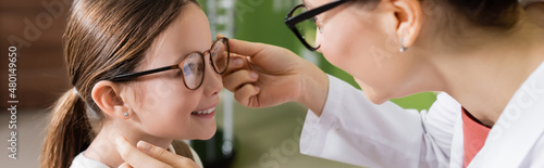 ophthalmologist trying eyeglasses on smiling girl in optics store, banner.