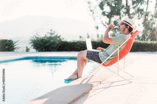 man sitting by the swimming pool with a laptop drinking beer alone