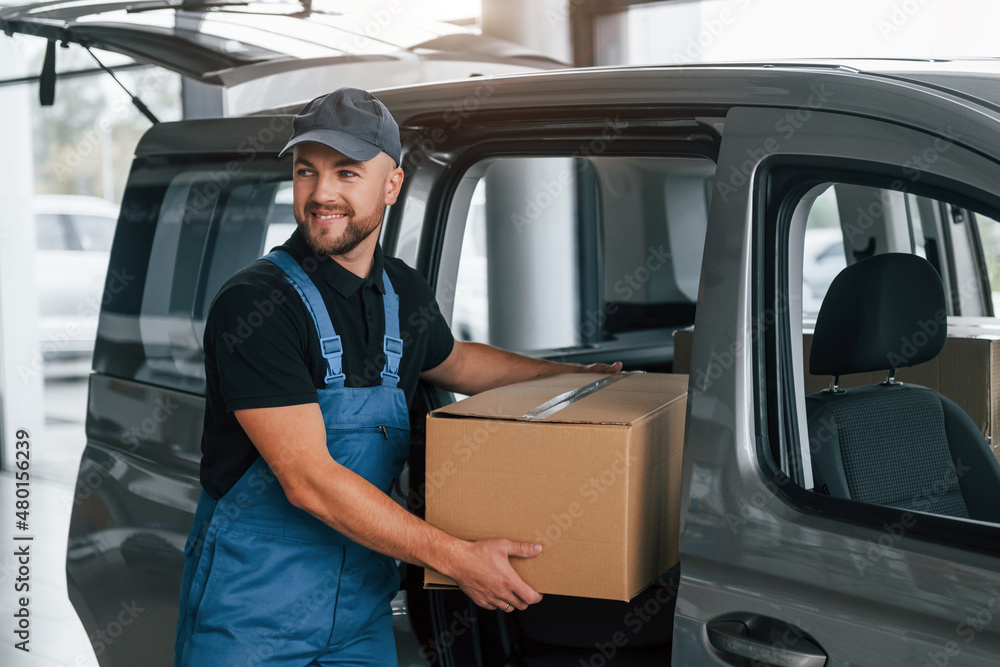With box in hands. Delivery man in uniform is indoors with car and with order With box in hands. Delivery man in uniform is indoors with car and with order