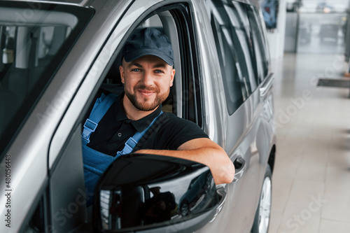 Sitting in vehicle and holding document. Delivery man in uniform is indoors with car and with order © standret