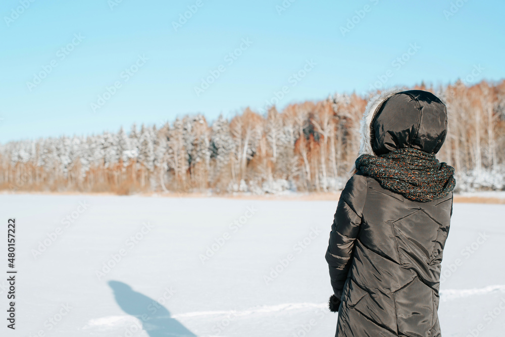 Back view of alone woman in hood and jacket standing on sunny frosty day in nature against backdrop of snowy forest and looking at winter landscape, copy space