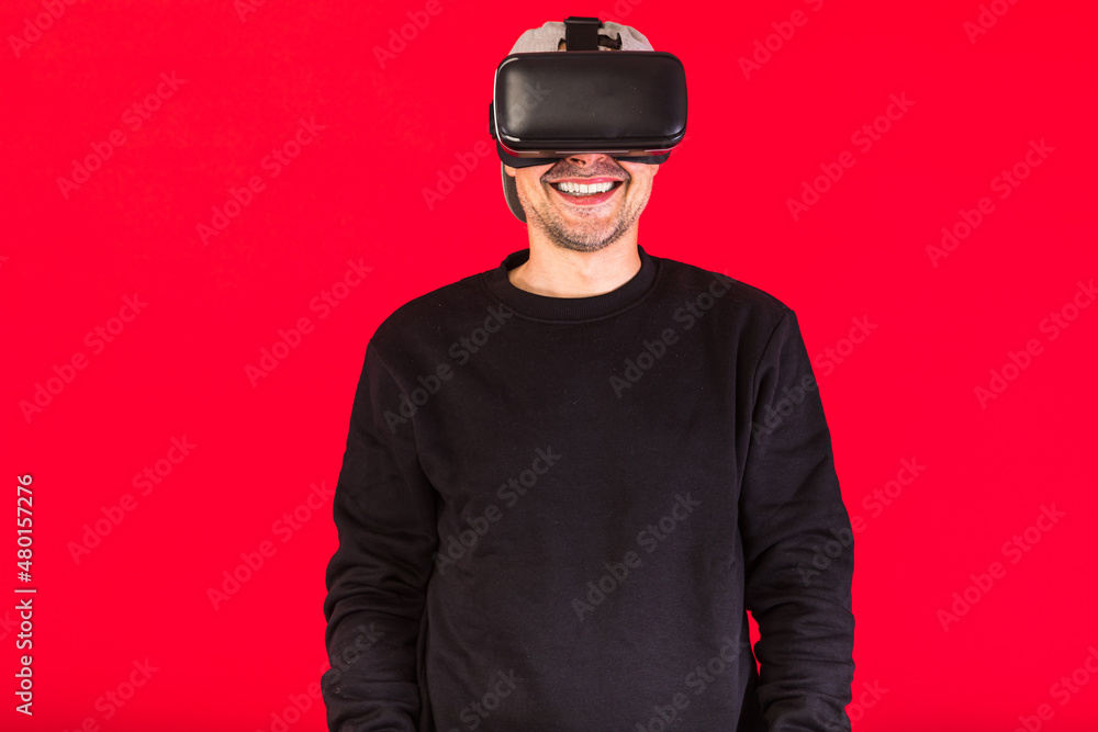 Man in cap dressed in black wearing Virtual Reality glasses, smiling, on red background. Technology, VR, computing and hobbies concept. Man in cap dressed in black wearing Virtual Reality glasses, smiling, on red background. Technology, VR, computing and hobbies concept.