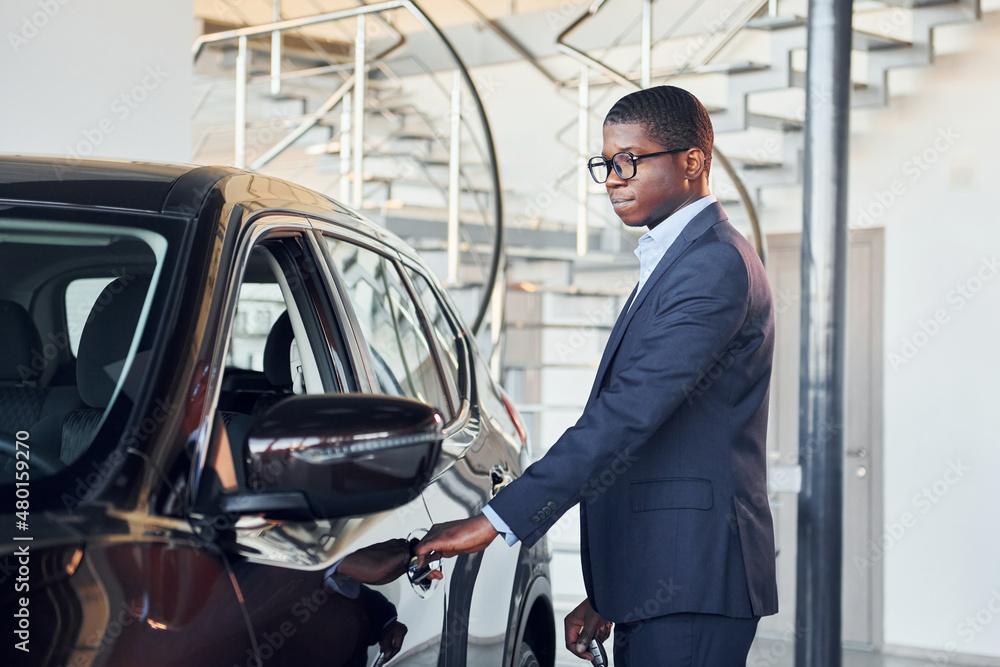 Opening the door. Young african american businessman in black suit is the autosalon