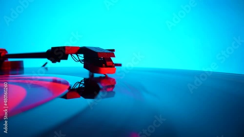 Black vinyl record spinning and play music on old turntable in rays of bright red and blue neon lights. Turntable needle on vinyl record, reads recording from grooves on disc. Close up. Slow motion.