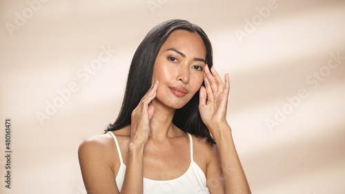 Female Beauty Portrait. Beautiful Asian Brunette Woman Posing, Smiling, Touching Her Face with Natural, Healthy Skin. Wellness and Skincare Concept on Soft Isolated Background. Close Up.