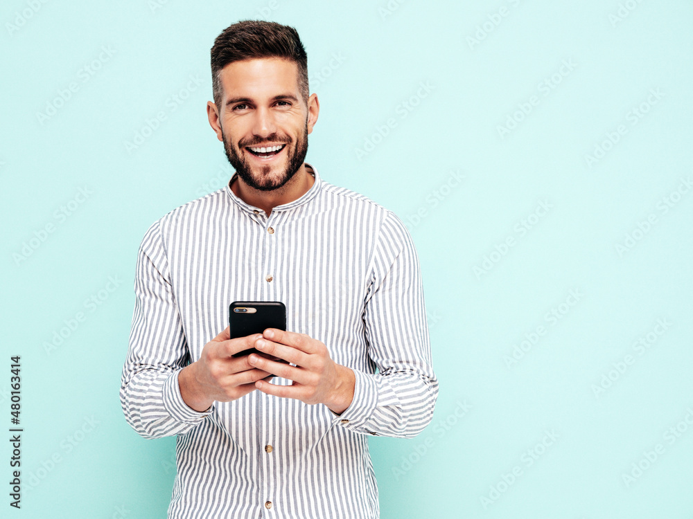 Handsome smiling model.Sexy stylish man dressed in shirt and jeans. Fashion hipster male posing near blue wall in studio. Holding smartphone. Looking at cellphone screen. Using apps Handsome smiling model.Sexy stylish man dressed in shirt and jeans. Fashion hipster male posing near blue wall in studio. Holding smartphone. Looking at cellphone screen. Using apps