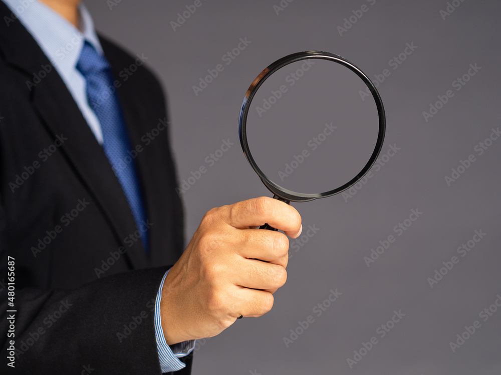 A businessman in a suit holding a magnifying glass while standing over a gray background A businessman in a suit holding a magnifying glass while standing over a gray background