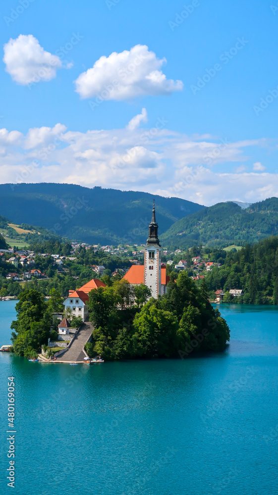 View of Catholic church situated on an island on Bled lake with mountains and villages on the background in the slovenian Alps