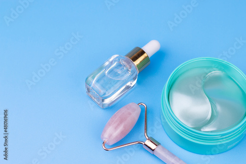 Face massage roller, hyaluronic patches and serum or oil on blue background. Collagen eye patches and face roller, flat lay, top view image. Skin care and spa concept.