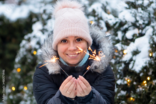 A happy woman holds a burning sparkler in her hand against the background of a Christmas tree, close-up, selective focus. Concept: festive mood, Christmas and New Year, joyful holiday. photo