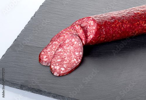A stick of smoked sausage and slices of sausage on a black cutting board