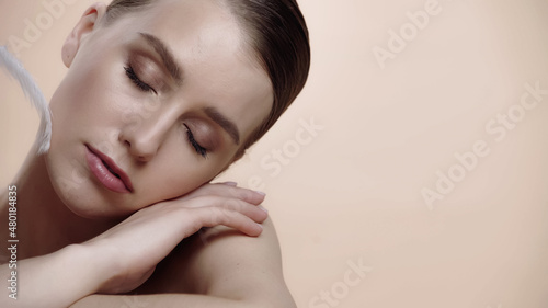 feather near cheek of pretty young woman with closed eyes isolated on beige.