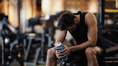 sportsman in earphone listening music, resting and holding sports bottle in gym.