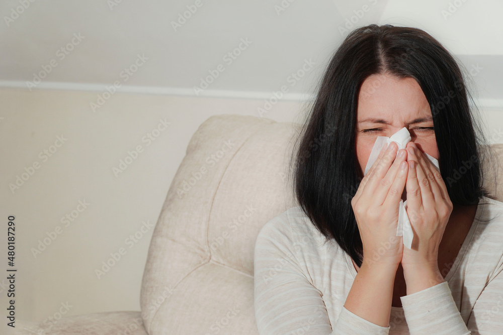 Middle aged mature woman suffering from allergy rhinitis at home. Symptoms of cold or allergy. Woman sneezes in white tissue. Middle aged mature woman suffering from allergy rhinitis at home. Symptoms of cold or allergy. Woman sneezes in white tissue.