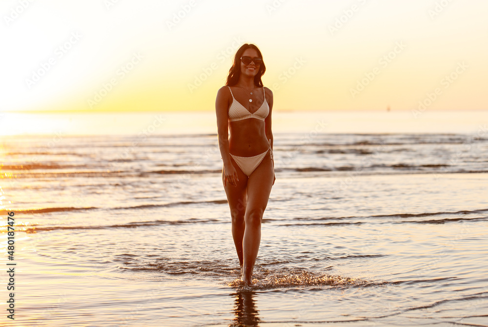 people, summer and leisure concept - happy smiling young woman in bikini  swimsuit on beach over sunset Stock Photo