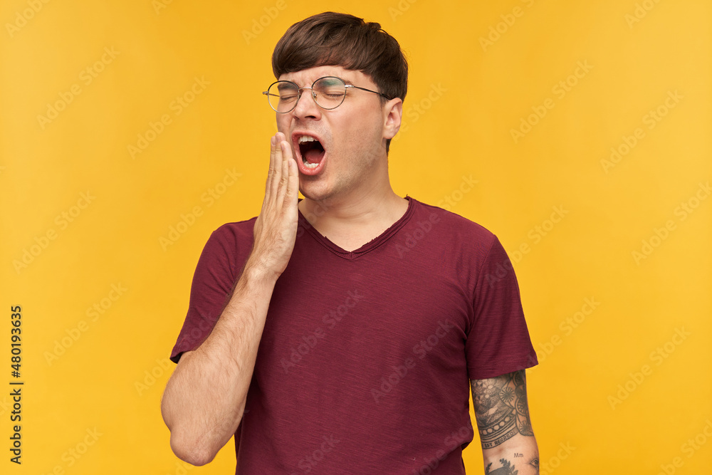 Studio shot of young tattooed brunette male, wears t-shirt and round glasses yawing, feels tired, isolated over yellow background Studio shot of young tattooed brunette male, wears t-shirt and round glasses yawing, feels tired, isolated over yellow background