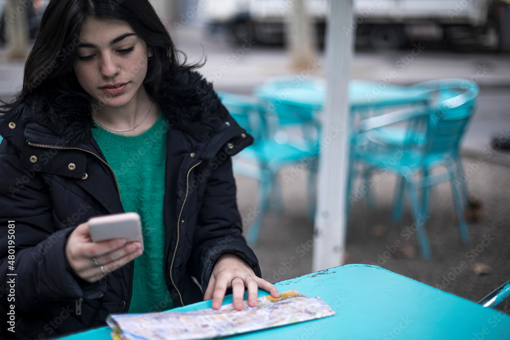 Fototapeta premium Young woman tourist is sitting in street cafe at table, looking for a way on map, using smartphone.