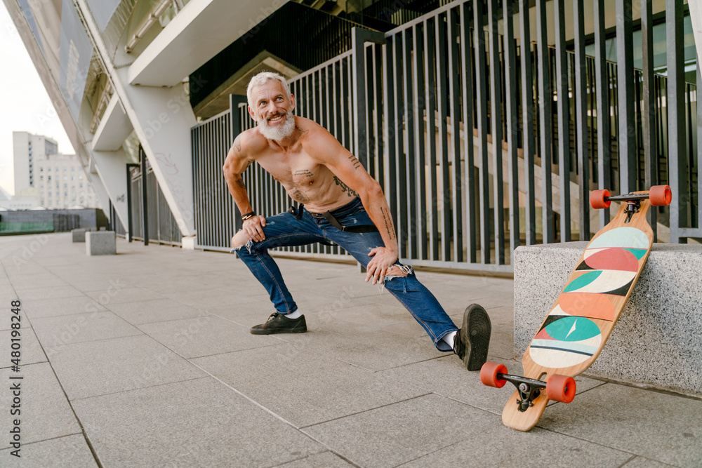 Adult white shirtless man doing workout by his skateboard outdoors