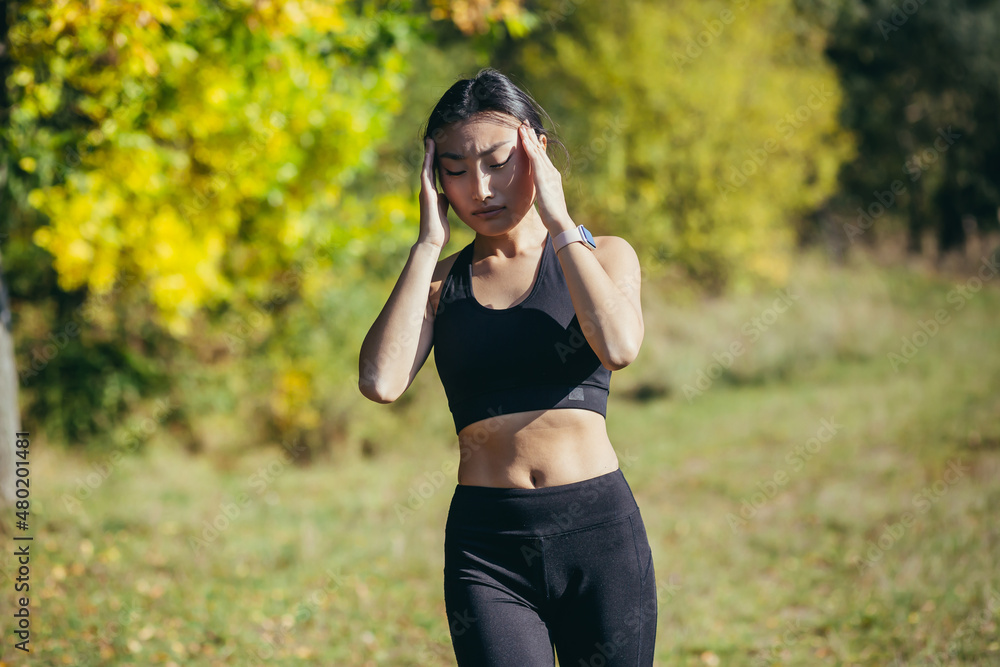A female athlete runs in the park, has a severe headache, Asian woman holds her hands behind her head A female athlete runs in the park, has a severe headache, Asian woman holds her hands behind her head