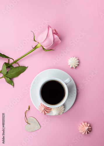 Black aromatic coffee in a white cup on a pink table. White and pink meringue cookies next to the cup. Pink rose in the background. Top view